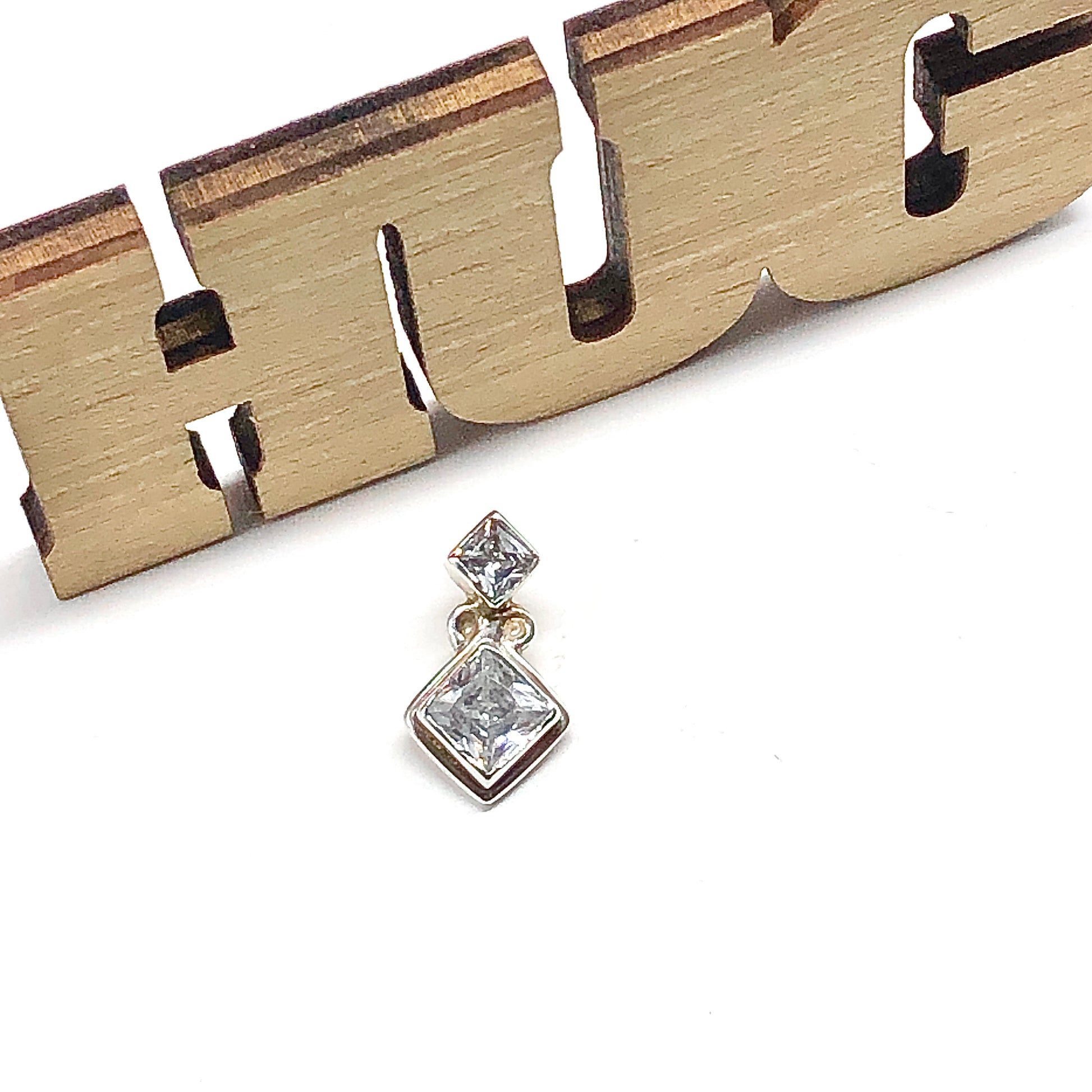 925 Silver 2 stone style Pendant with a clear cz gemstone in front of a wooden cutout spelling 'HUG' on a white table image