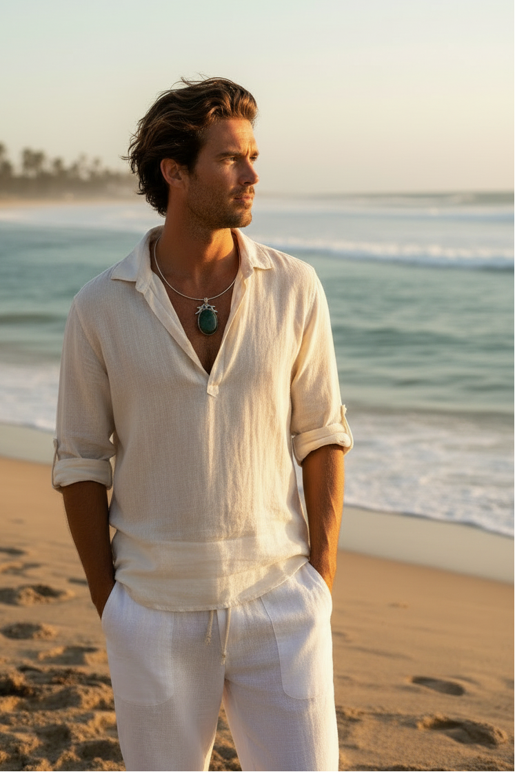 A man standing on a sandy beach near sunset, wearing a loose white linen shirt large green stone pendant necklace and white pants, with the ocean and palm trees in the background.