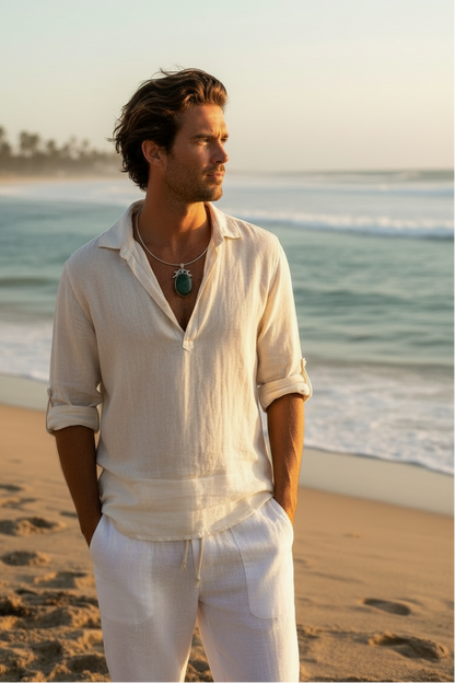 A man standing on a sandy beach near sunset, wearing a loose white linen shirt large green stone pendant necklace and white pants, with the ocean and palm trees in the background.